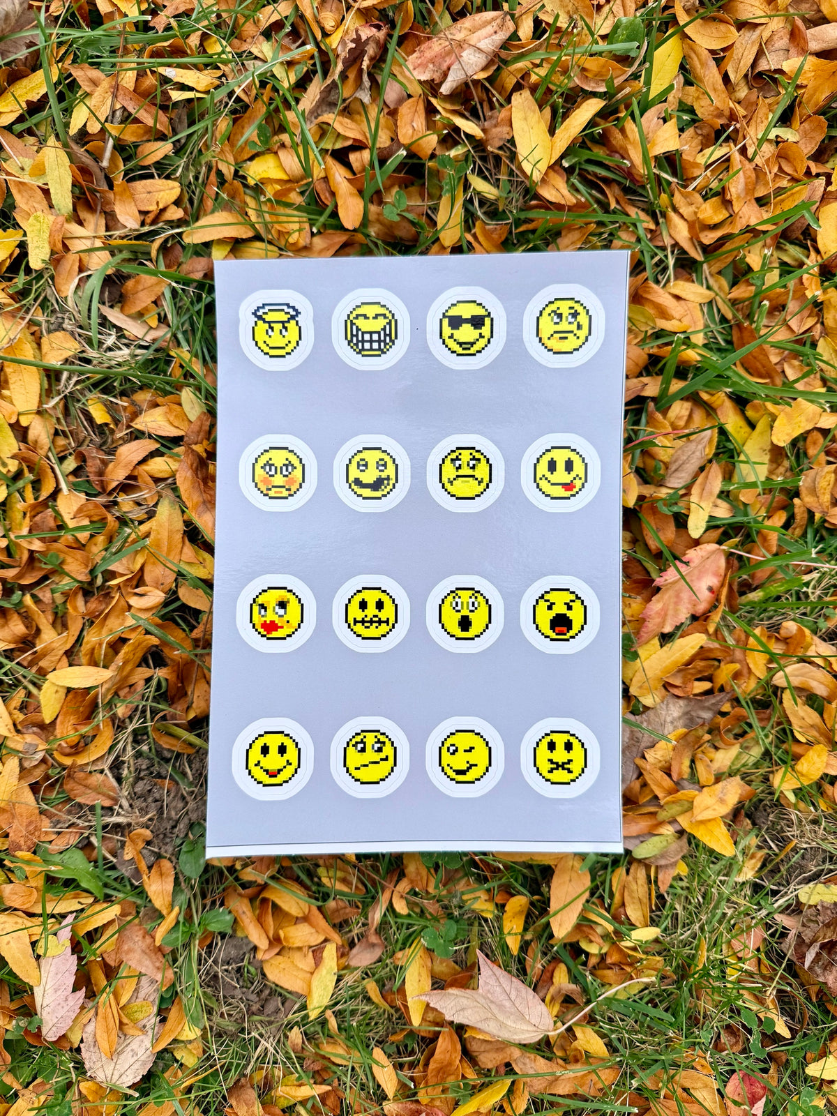 AIM Emoticon Sticker Sheet on fall leaves: 16 round pixel-art yellow emoticon stickers on a gray backing sheet